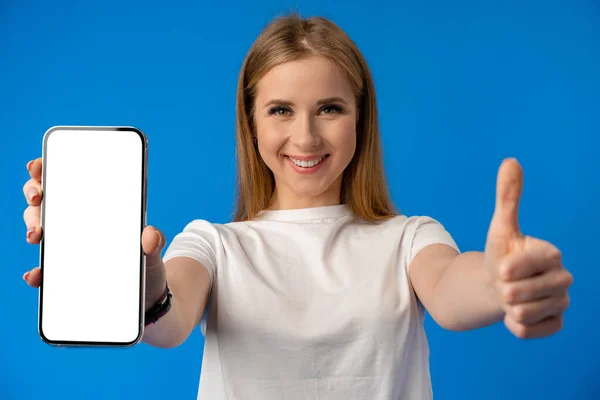 Mujer joven señala con el dedo la pantalla aislada del teléfono inteligente sobre fondo de color azul — Foto de Stock