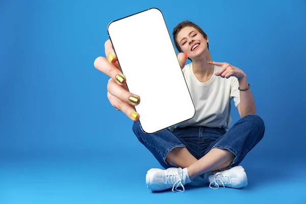 Mujer feliz muestra pantalla de teléfono inteligente en blanco contra fondo azul — Foto de Stock