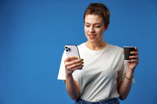 Jovem atraente alegre segurando smartphone, desfrutando de bate-papo ou compras on-line contra fundo azul — Fotografia de Stock