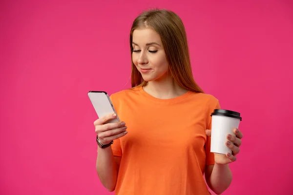 Jovem mulher usando smartphone e segurando xícara de café sobre fundo rosa — Fotografia de Stock
