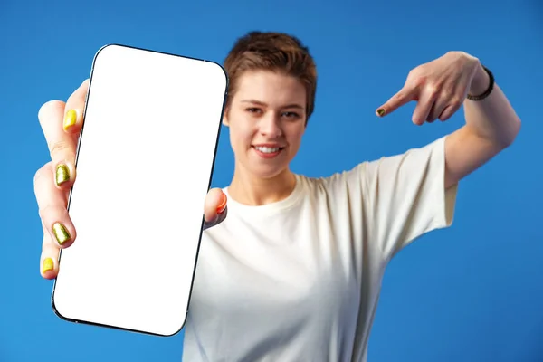 Retrato de una mujer joven mostrando el teléfono móvil en blanco mientras está de pie sobre el fondo azul — Foto de Stock