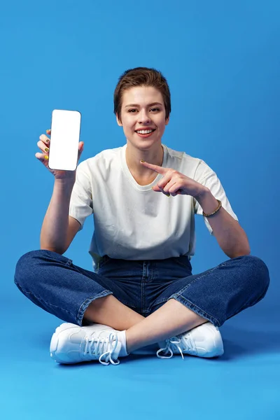 Glückliche Frau zeigt leeren Smartphone-Bildschirm vor blauem Hintergrund — Stockfoto