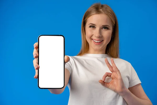 Młoda kobieta wskazuje palcem na pojedynczy ekran smartfona na tle niebieskiego koloru — Zdjęcie stockowe