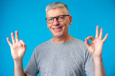 Mutlu pozitif yakışıklı yaşlı adam mavi arka planda tamam işareti gösteriyor.
