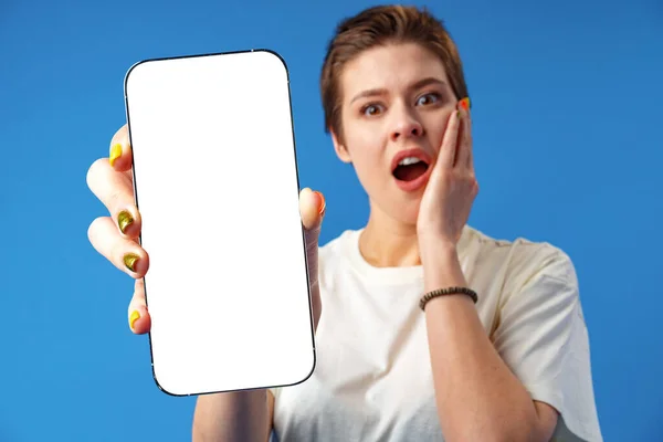Retrato de uma jovem que mostra o telefone celular em branco da tela enquanto está em cima do fundo azul — Fotografia de Stock