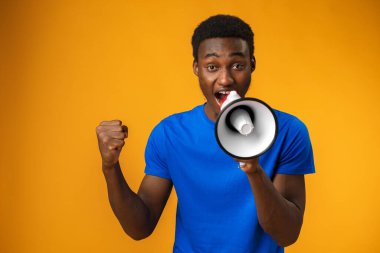 Genç siyah adam sarı arka planda megafonla bağırıyor.