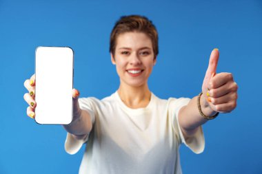 Mavi arka planda dikilirken boş ekran cep telefonu gösteren genç bir kadının portresi