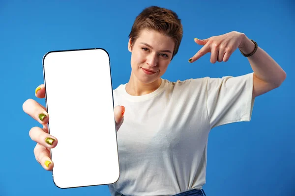 青い背景の上に立っている間、空白の画面の携帯電話を示す若い女性の肖像画 — ストック写真