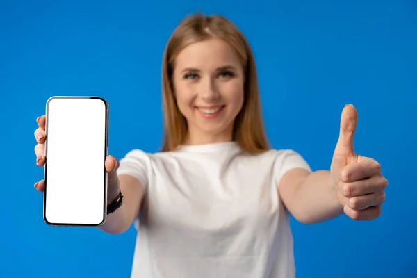 Mujer joven señala con el dedo la pantalla aislada del teléfono inteligente sobre fondo de color azul — Foto de Stock