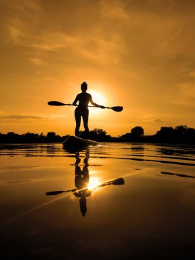 Yemek masasında bir kadın, gün batımında sörf yaparken suda yansıyor.