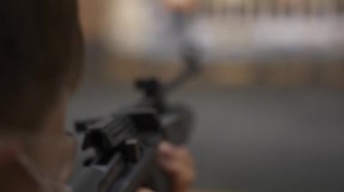 Bir çocuk siyah havalı tüfekten nişan alır. Parçalama gözlüğü ve tıbbi maske takar.