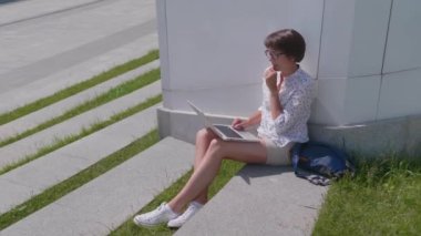 Gözlüklü iş kadını şehir parkında dizüstü bilgisayarla çalışıyor. Yazın serbest çalışanlar için açık hava işyeri.