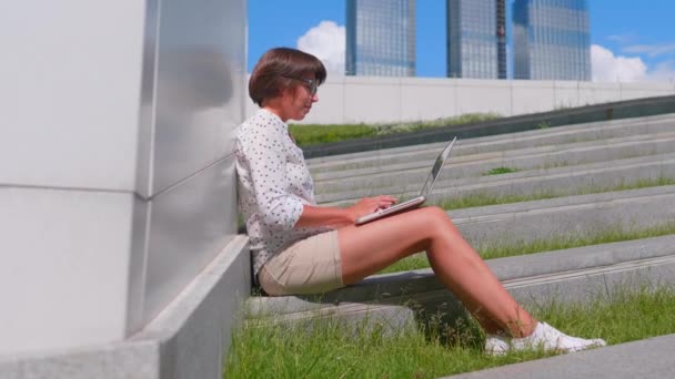 Egy szemüveges üzletasszony laptoppal dolgozik a városi parkban. Szabadúszók szabadtéri munkahelye nyáron. — Stock videók