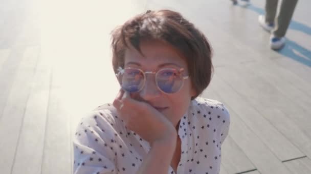Donna con occhiali da sole colorati siede sulla scena nel parco urbano e gode di luce solare brillante. Calore estivo in città. — Video Stock