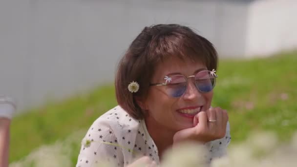 다채 로운 선글라스를 쓴 여자는 도시 공원의 잔디밭에 누워 꽃을 즐긴다. 마을의 여름 더위. — 비디오
