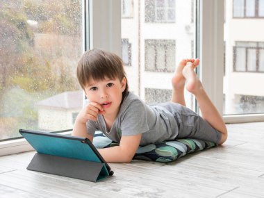 Meraklı çocuk dijital tablette çizgi film izliyor. Çocuk yerde oturuyor ve elektronik cihaz kullanıyor. Dışarıda yağmur yağarken çocuklar için kapalı bir eğlence..