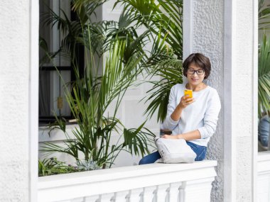 Akıllı telefondan mesaj atan bir kadın. Gözlüklü güzel bir kadın çiçek saksılarında palmiye ağaçlarıyla balkonda oturuyor. Tropik ev bitkisi. Yaz titreşimleri.