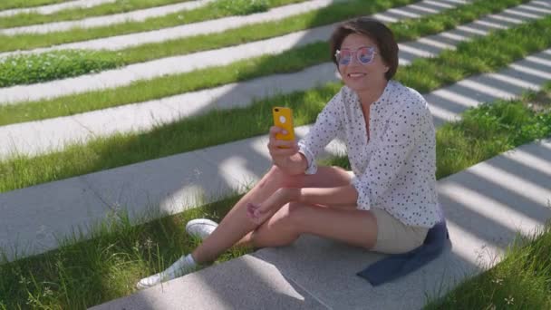 Woman with colorful sunglasses sits on lawn in urban park and makes selfie on smartphone. Summer heat in town. — Stock Video