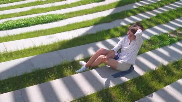 都会の公園の芝生の上にカラフルなサングラスの女性が座り、スマートフォンで自撮りをします。町の夏の暑さ. — ストック動画