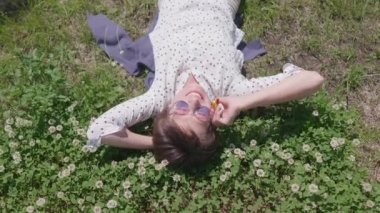 Şehir parkında çimlere uzanmış renkli güneş gözlüklü bir kadının manzarası ve çiçeklerden hoşlanıyor. Kasabada yaz sıcağı.
