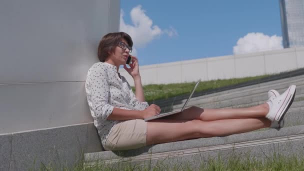 Жінка в окулярах працює з ноутбуком-мурахами на смартфоні в міському парку. На відкритому повітрі для фрілансерів влітку . — стокове відео