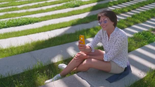 Woman with colorful sunglasses sits on lawn in urban park and makes selfie on smartphone. Summer heat in town. — Stock Video