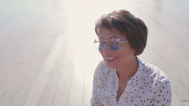 Woman with colorful sunglasses sits on scene in urban park and enjoys of bright sunlight. Summer heat in town. — Stock Video