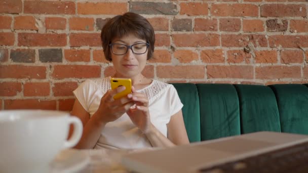 Жінка працює дистанційно в кафе з стінами з червоної цегли. Жінка в окулярах прокручує екран смартфона. Співпрацюючий центр з стилем інтер'єру лофт. позаштатне робоче місце . — стокове відео