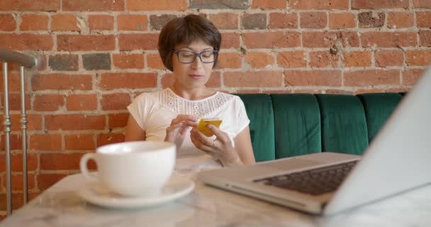 Kobieta pracuje zdalnie w kawiarni z czerwoną ceglaną ścianą. Kobieta w okularach rozmawia przez smartfona. Współpracujące centrum ze stylem wnętrz poddasza. Miejsce pracy na własny rachunek. — Wideo stockowe