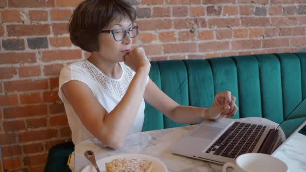 Kvinna arbetar på distans i café med röda tegelväggar. Kvinna i glasögon typer på laptop tangentbord, dricker kaffe och äter tårta. Co-working center med loft interiör stil. Frilansande arbetsplats. — Stockvideo