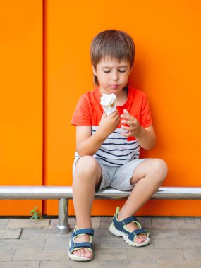 Küçük çocuk dışarıda dondurma yiyor. Tek renkli çocuk, parlak turuncu duvar arkaplanı. Yazın lezzetli soğuk bir tatlı..