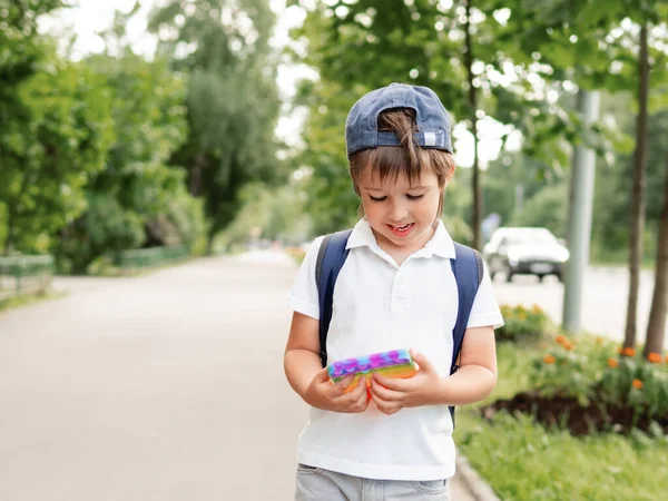 Little boy plays square rubber fidget toy Pop It. Rainbow-colored anti stress and tactile toy for all ages. Outdoor recreation with toy for fine motor skills improvement.