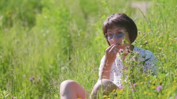 Женщина в красочных солнцезащитных очках наслаждается солнечным светом и цветочным ароматом на травяном поле. Летние вибрации. Расслабься на улице. Самоуспокоение. — стоковое видео