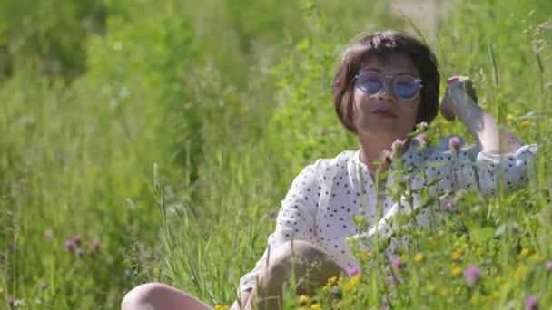 Kvinna i färgglada solglasögon njuter av solljus och blomdoft på gräsplan. Sommarvibbar. Slappna av utomhus. Självlindrande. — Stockvideo