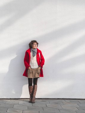 Kırmızı spor ceketli kıvırcık kadın güneş ışınlarıyla beyaz duvarın yanında duruyor. Gülümseyen öğrenci. Modern yaşam tarzı.