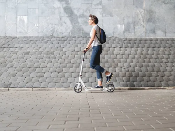 女性はキックスクーターに乗る ジーンズとスニーカーの女性は灰色のタイル張りの壁の背景で速く動きます 健康的なライフスタイル 環境に優しい都市交通 — ストック写真