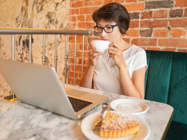 Woman works remotely in cafe with red brick walls. Female in eyeglasses types on laptop keyboard, drinks coffee and eats a tart. Co-working center with loft interior style. Freelance workplace.