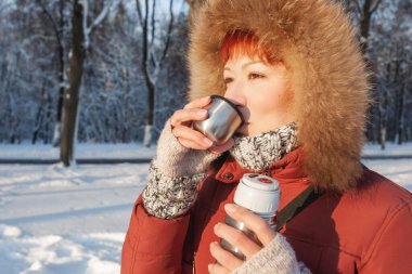 Kırmızı ceketli kadın metal termostan sıcak çay içiyor. Günbatımında kış ormanlarında boş zaman aktivitesi.