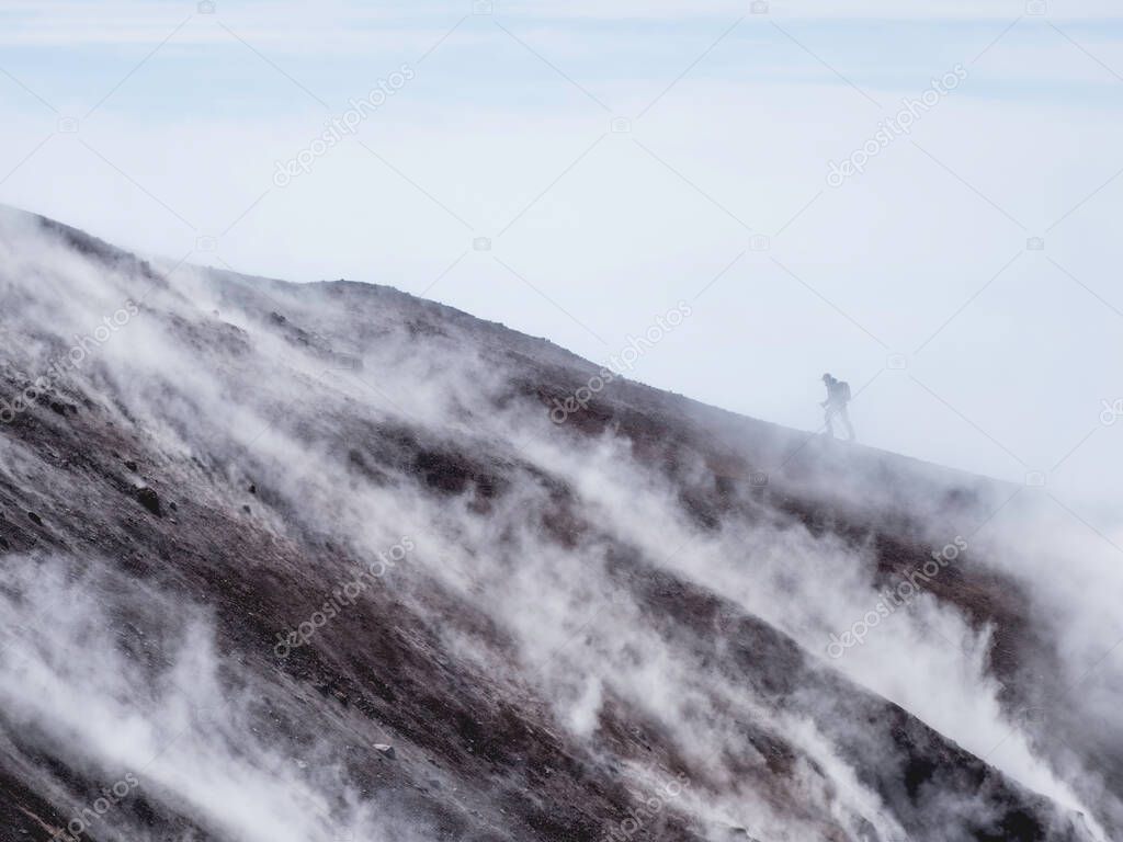 Silhouette of man hiking at coldera of Avachinsky stratovolcano, also known as Avacha Volcano. Backpacker tourist moves upon rocks behind steam from hot geysers. Kamchatka Peninsula, Russia.