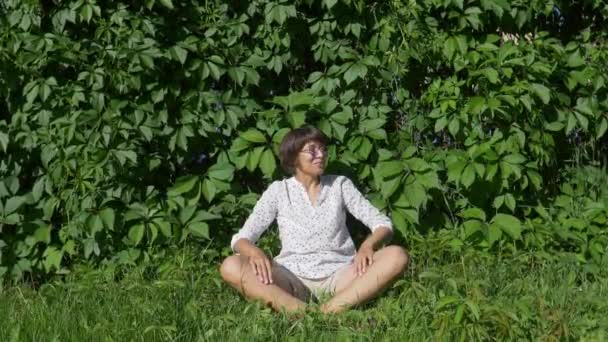 Brett leende kvinna sitter på gräsmattan nära väggen täckt med tjock murgröna. Naturlig bakgrund med gröna blad och kvinna njuter av solljus och värme i stadsparken. Sommarstämningar. — Stockvideo