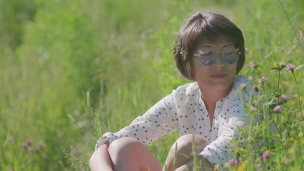 Kvinna i färgglada solglasögon njuter av solljus och blomdoft på gräsplan. Sommarvibbar. Slappna av utomhus. Självlindrande. — Stockvideo