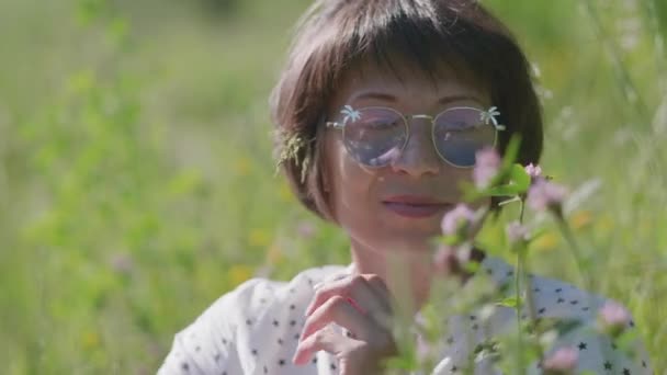 Kvinna i färgglada solglasögon njuter av solljus och blomdoft på gräsplan. Sommarvibbar. Slappna av utomhus. Självlindrande. — Stockvideo