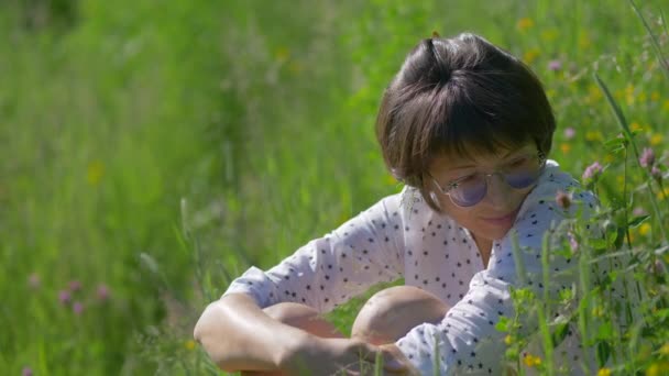 Kvinna i färgglada solglasögon njuter av solljus och blomdoft på gräsplan. Sommarvibbar. Slappna av utomhus. Självlindrande. — Stockvideo