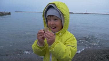 Sarı tulumlu küçük çocuk Karadeniz kıyısında oynuyor. Deniz sörfünün yanındaki çakıl taşlı sahilde oynaşan bir çocuk..