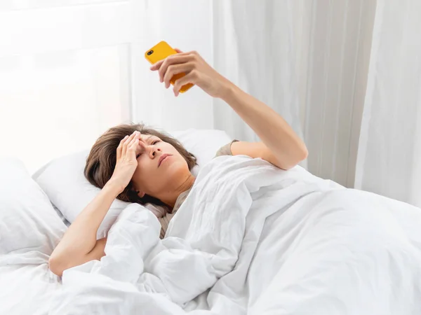 眠い女はスマートフォンの画面を見ている 目が覚めた直後にメールボックスをチェックします ベッドで無線技術を使う — ストック写真