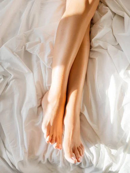 Morning in bed. Woman\'s feet on unmade bed. Sunbeams on white crumpled bed sheet. Light and shadow.
