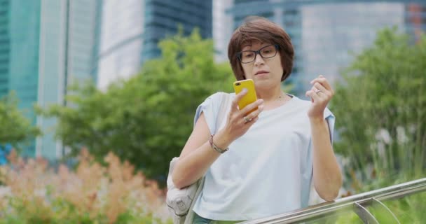 Kvinnan talar via smartphone. Söt kvinna i glasögon talar i urban park på modern byggnad bakgrund. — Stockvideo