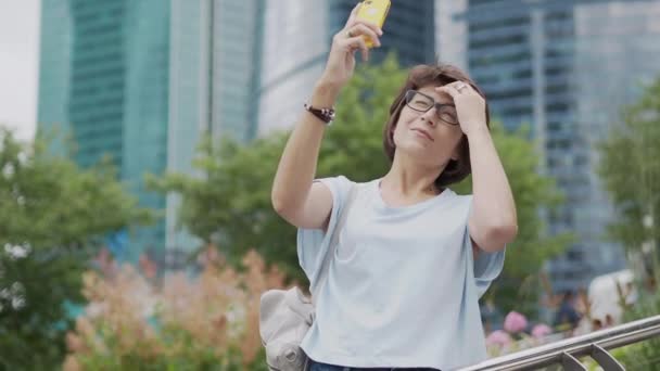 Woman makes selfie on smartphone. Pretty female in eyeglassesin urban park at summer. Vodern building on background. City life in big town. — Stock Video