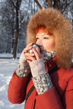 Kırmızı ceketli kadın metal termostan sıcak çay içiyor. Günbatımında kış ormanlarında boş zaman aktivitesi.