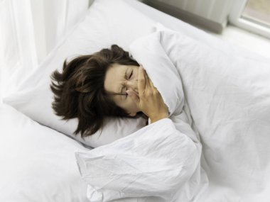 Hasta kadın yatakta öksürüyor. Açık renkli yatak odası ve beyaz çarşaflar. Coronavirus COVID19 karantinası. Evde kendini izole et. Evde sabahlar. 